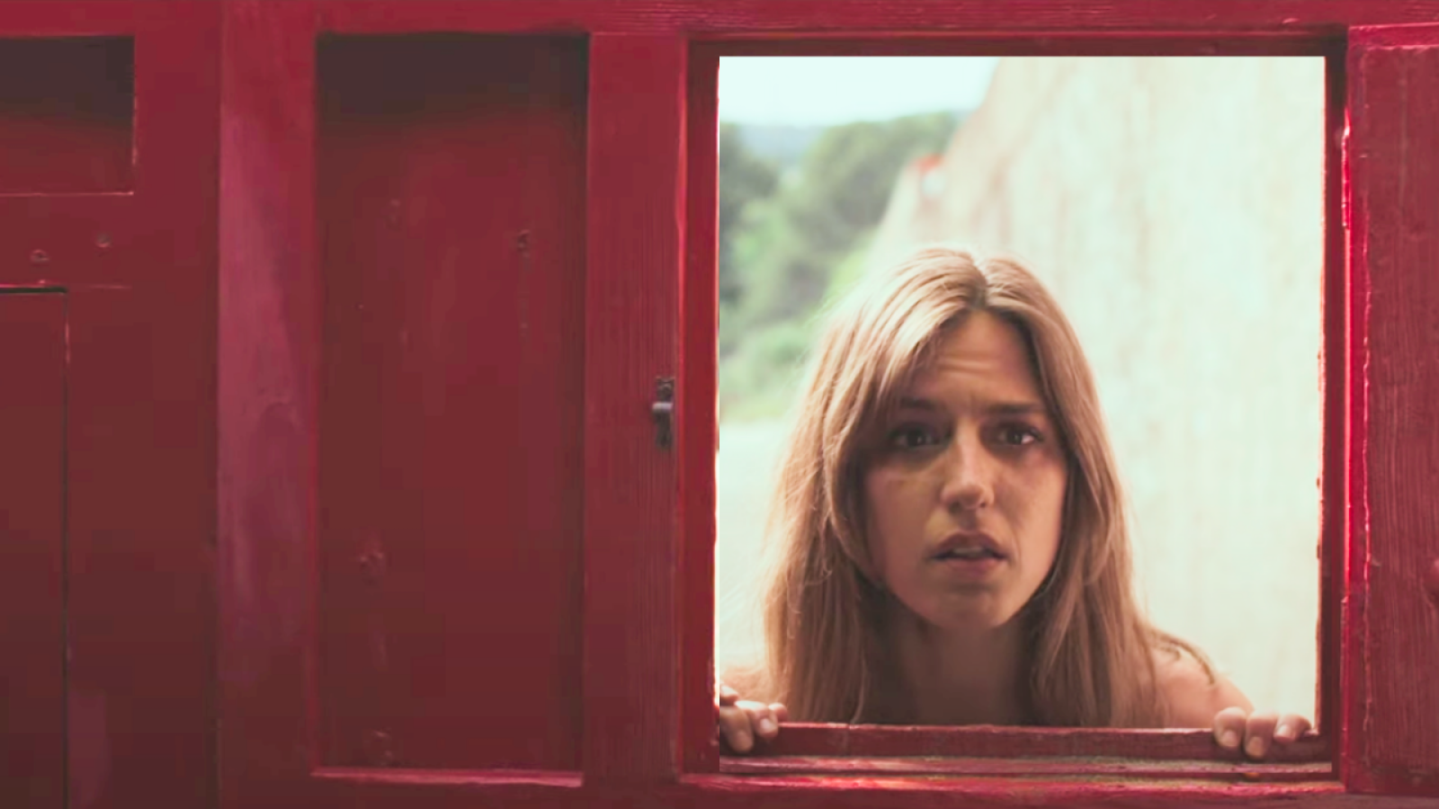 Actriz con expresión seria mirando a través de una ventana con marco rojo; fondo borroso con vegetación.