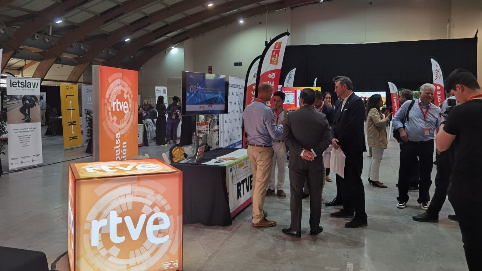 Stand de RTVE en un evento, con logo naranja y blanco, pantallas y varias personas en vestimenta formal. Se muestra información sobre un proyecto.