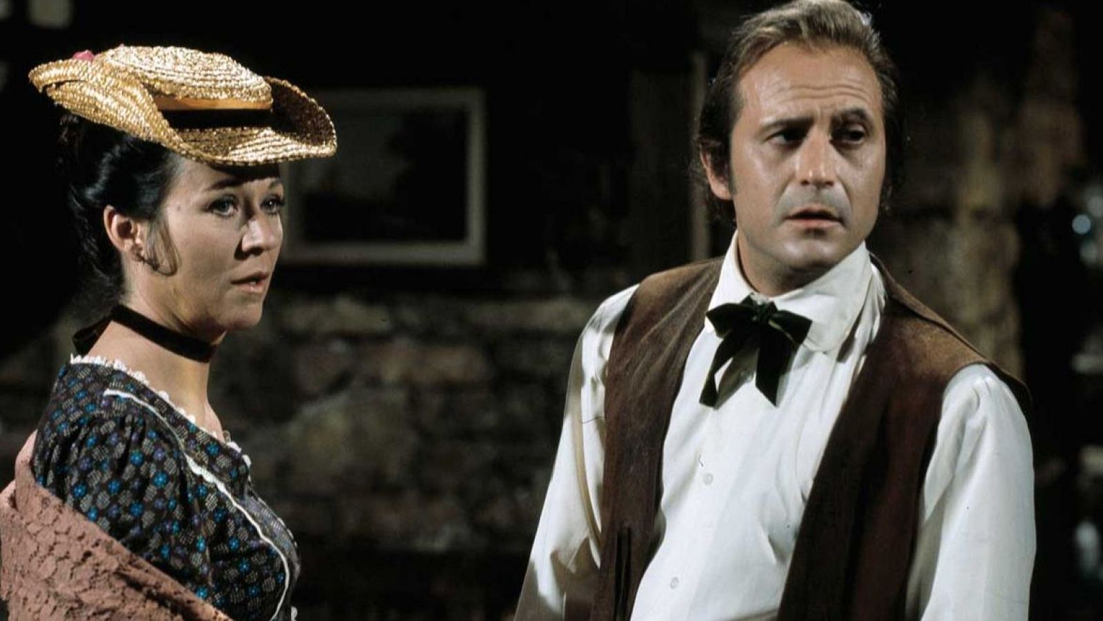 Julieta Serrano con sombrero de paja y vestido oscuro, y Agustín González con camisa blanca y chaleco marrón, en una escena de interior con iluminación tenue.