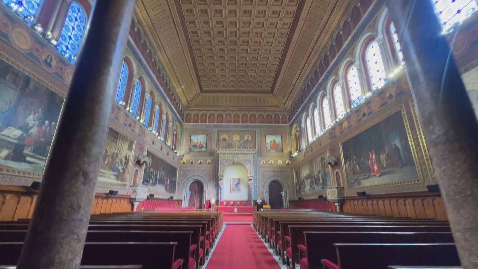 Imponente auditorio con techo artesonado, paredes con pinturas murales, arcos, columnas y bancos de madera dispuestos simétricamente alrededor de una alfombra roja.