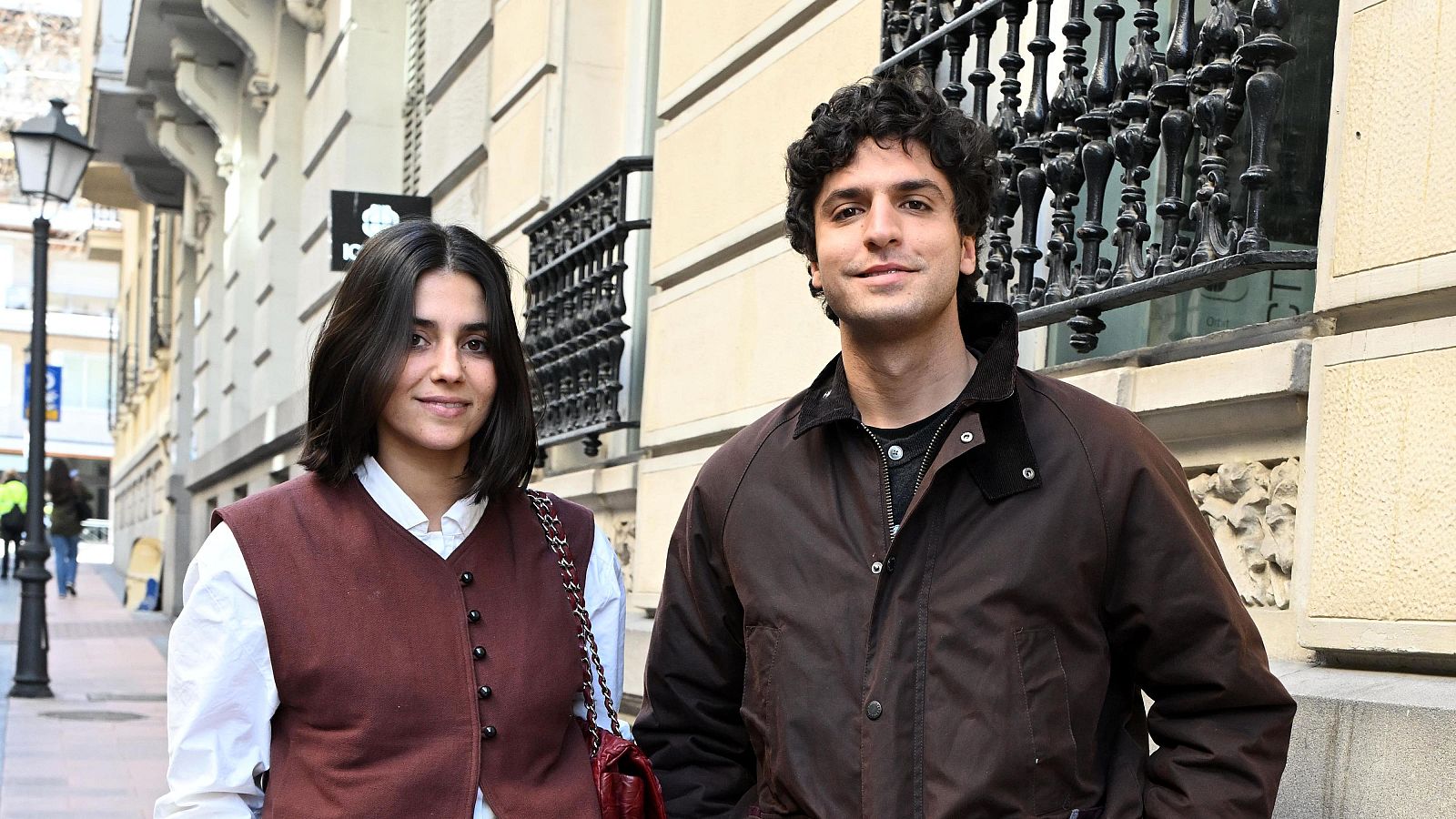 Dos personas, una mujer con camisa blanca y chaleco marrón, y un hombre con chaqueta marrón oscura, posan juntas en una calle frente a un edificio claro con detalles arquitectónicos.