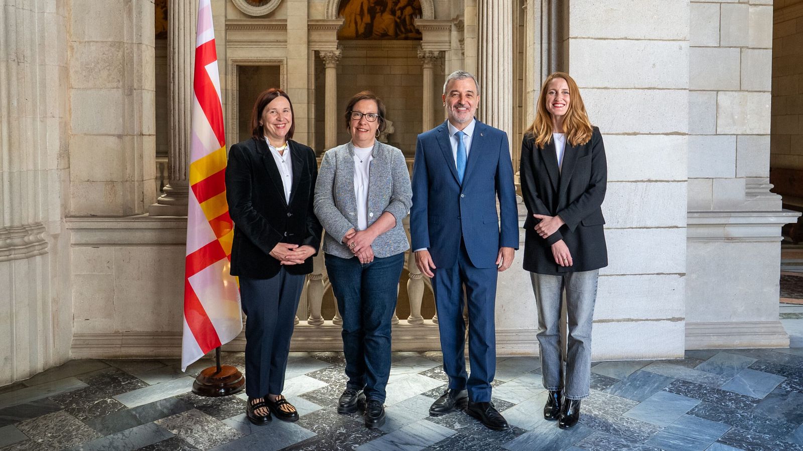 Cuatro personas, dos hombres y dos mujeres en atuendo formal, posan para una foto en un interior con columnas y una bandera.  Contexto político municipal.