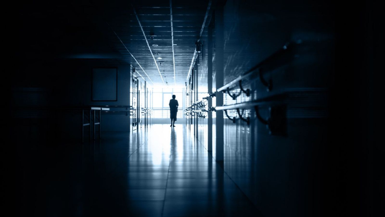 Silueta caminando por un pasillo de hospital oscuro hacia una ventana iluminada. Barandillas en las paredes, atmósfera sombría con un toque de esperanza.