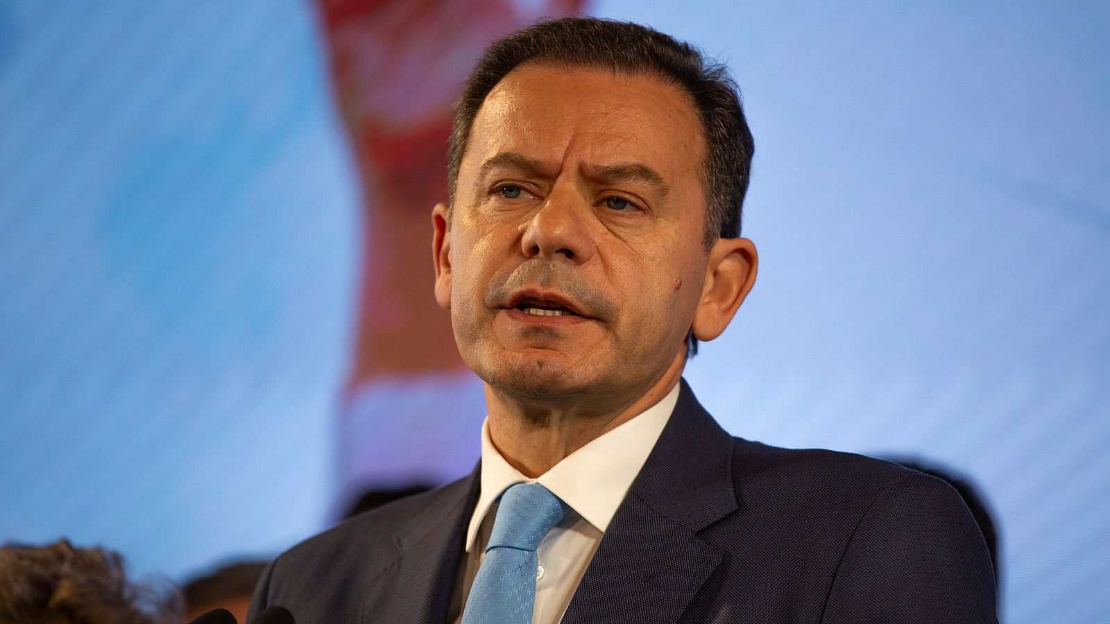 Ex primer ministro montenegrino, en traje oscuro y corbata azul, habla ante micrófonos en un evento con fondo azul claro.