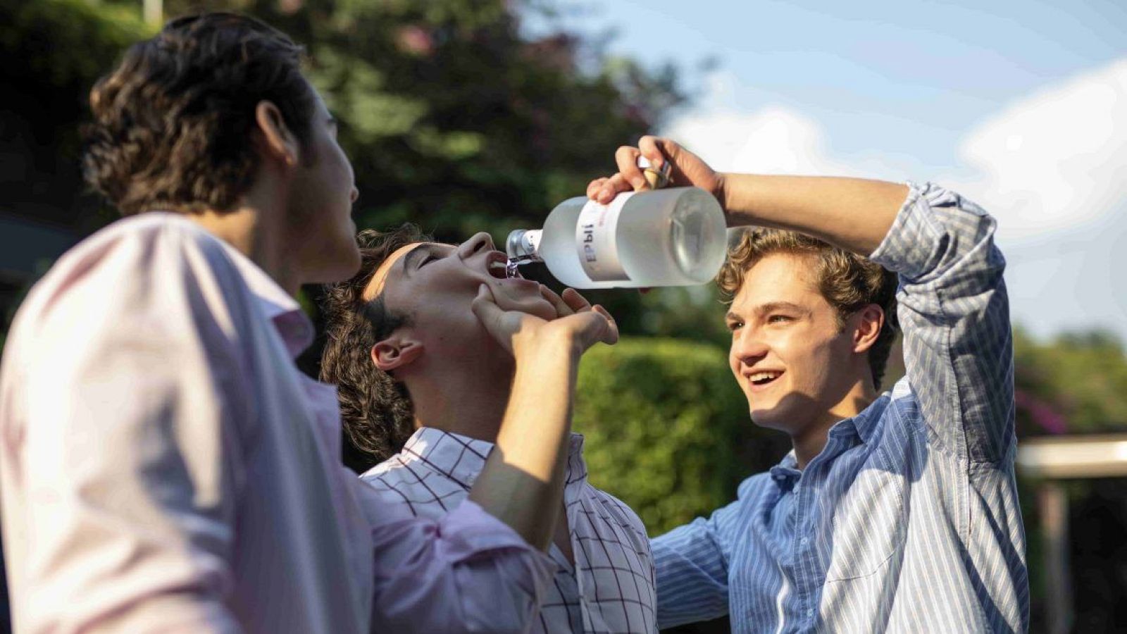 Jóvenes disfrutando en un entorno exterior; uno vierte líquido de una botella a otro, mientras un tercero observa.  Escena festiva con implicaciones negativas.