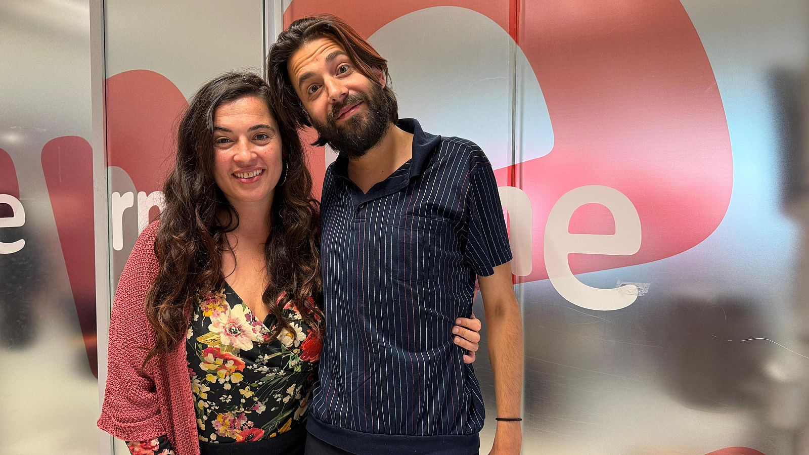 Dos cantantes, ella con chaqueta rosa y él con polo de rayas, posan sonrientes en un estudio de radio con el logo de RNE al fondo.