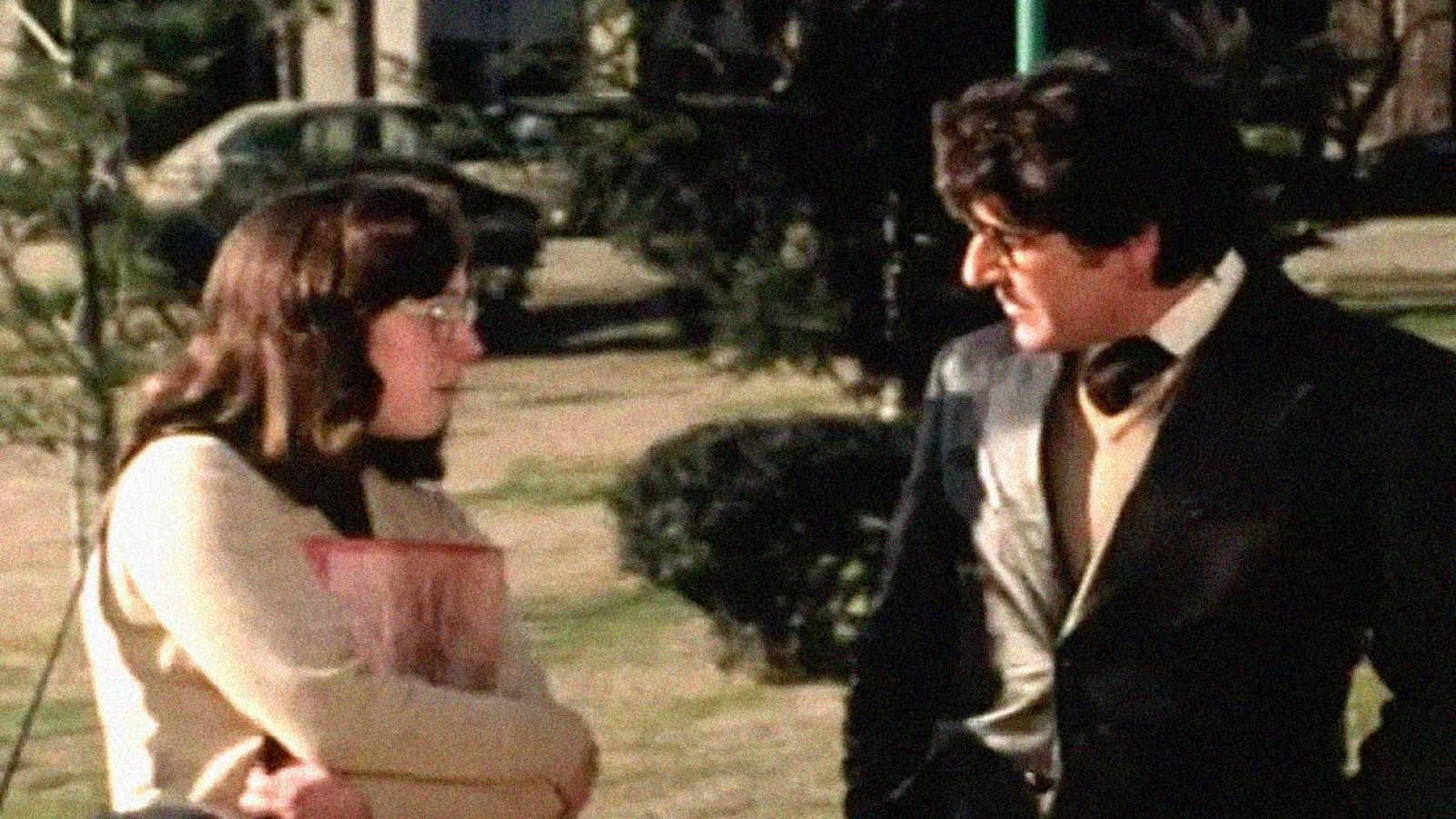 Escena exterior de entrevista en 'La España de los Botejara': Alfredo Amestoy, en traje oscuro y pajarita, conversa con una mujer con gafas que sostiene un objeto rectangular.