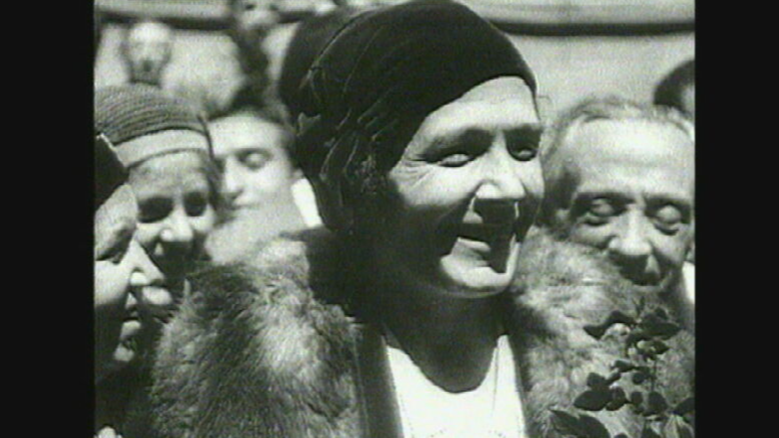 Victoria Kent en primer plano, con sombrero oscuro y expresión alegre, en una foto en blanco y negro de un evento público con otras personas.