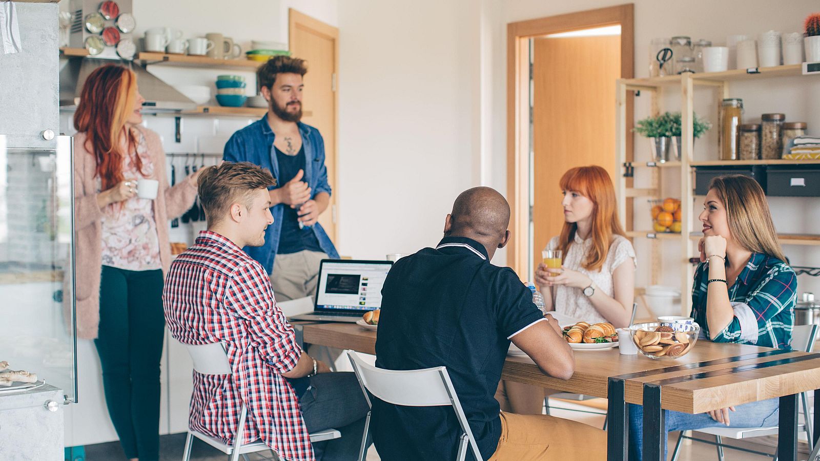 Seis personas conversan en una cocina, compartiendo comida y bebida en un ambiente informal y amigable, representativo de la vida en un co-living.
