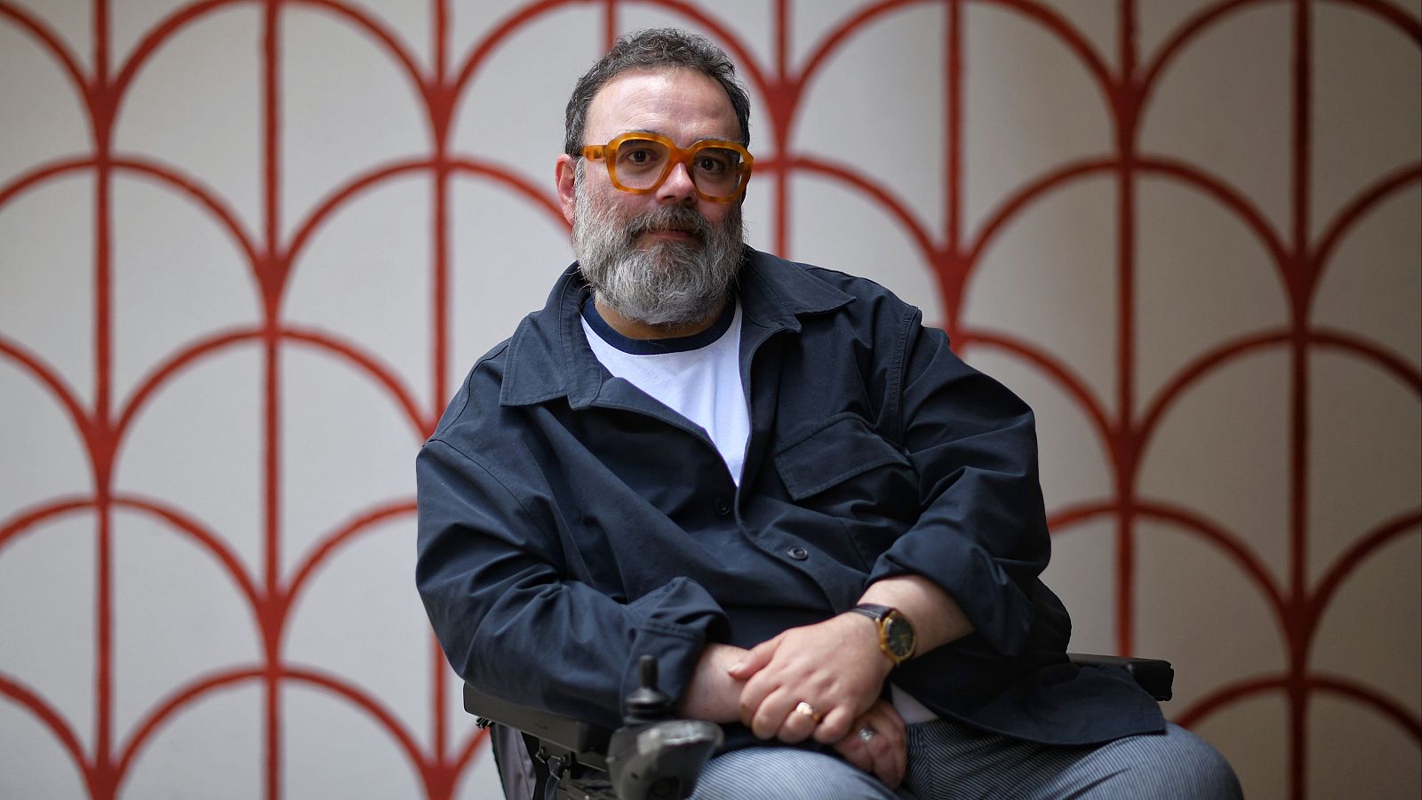 Retrato de Bob Pop: gafas ámbar, camisa azul sobre blanca, manos entrelazadas, reloj y anillo. Fondo geométrico rojo y blanco.