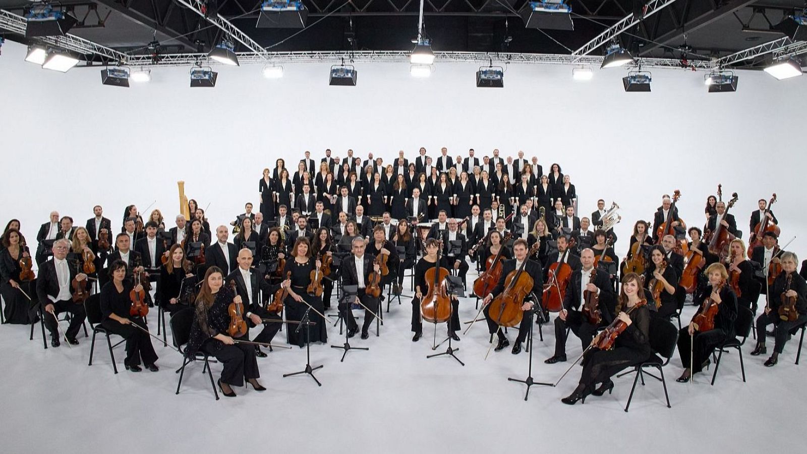 Orquesta y Coro RTVE posando en formación para una foto.  Músicos vestidos de negro con instrumentos de cuerda, viento, percusión y arpa. Fondo blanco de estudio.