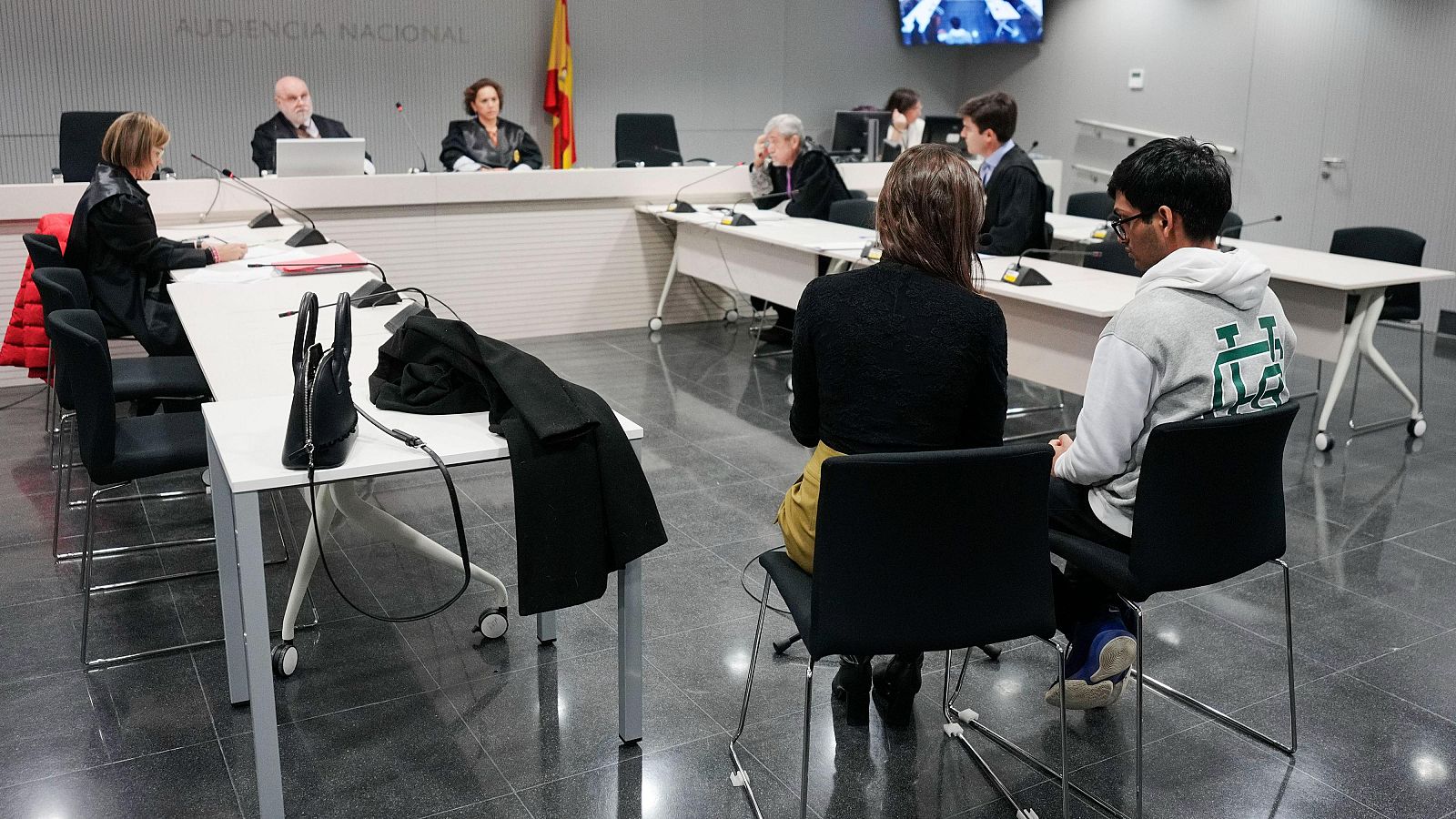 Sala de juicio con jueces, acusado y acompañante sentados, y otras personas presentes.  Se aprecia la bandera de España y el rótulo 'Audiencia Nacional'.