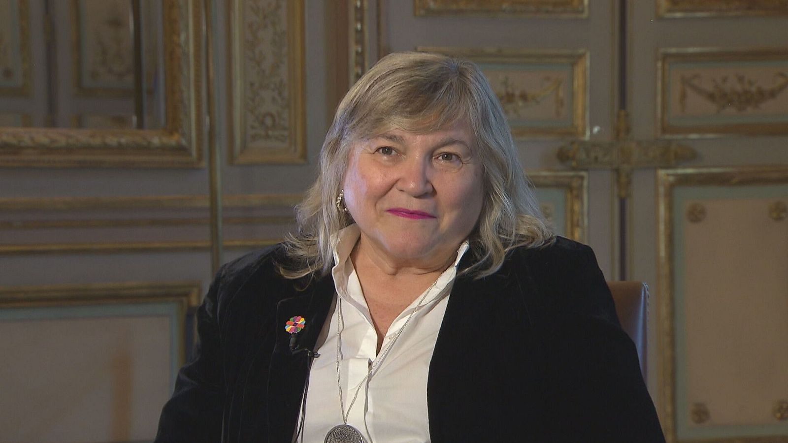 Mujer de edad madura con chaqueta oscura y blusa blanca, sentada en un interior elegante, posiblemente durante una entrevista. Lleva un collar con medallón y un pequeño dispositivo en la solapa.