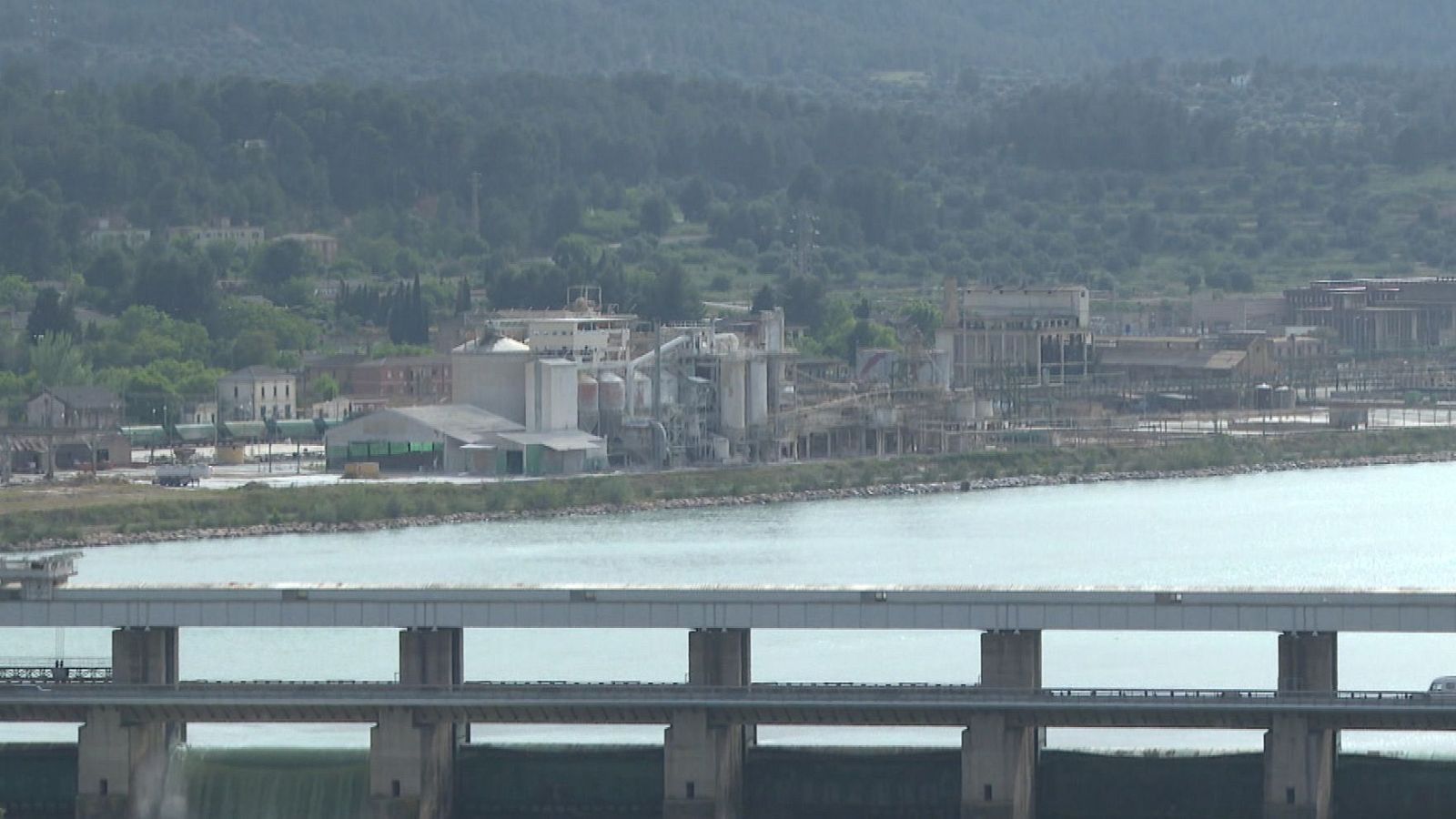 Vista panorámica de zona industrial junto a un río, con planta química, estructuras (posiblemente puente o presa), y vegetación de fondo.