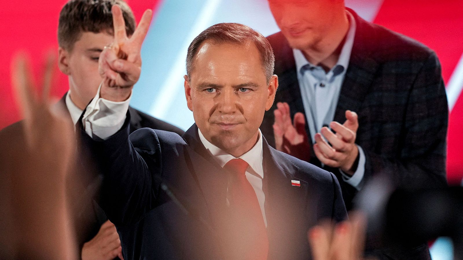 Candidato presidencial polaco haciendo el gesto de la victoria en un evento público. Viste traje oscuro y corbata roja, con una pequeña bandera en la solapa.