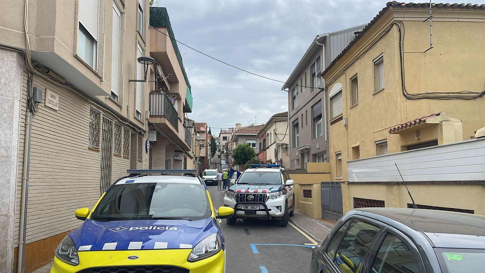 Escena policial en una calle con dos vehículos policiales, un cuerpo sin vida y una persona herida en un domicilio próximo.  Varios curiosos observan la situación.