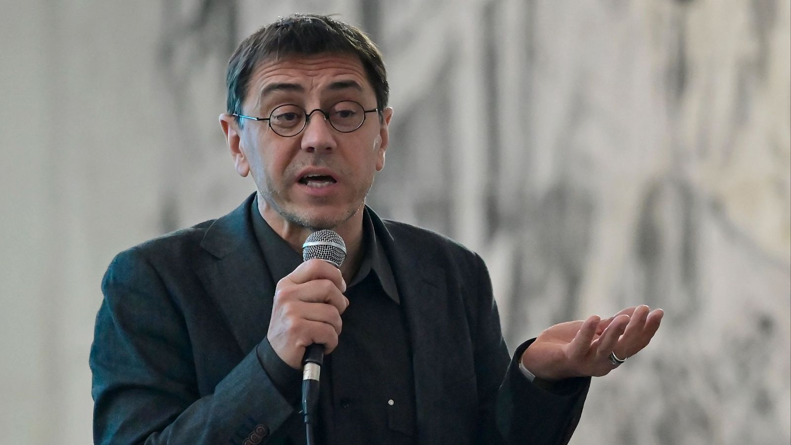Juan Carlos Monedero, en imagen, habla por micrófono; viste chaqueta oscura y camisa negra; el fondo está desenfocado.