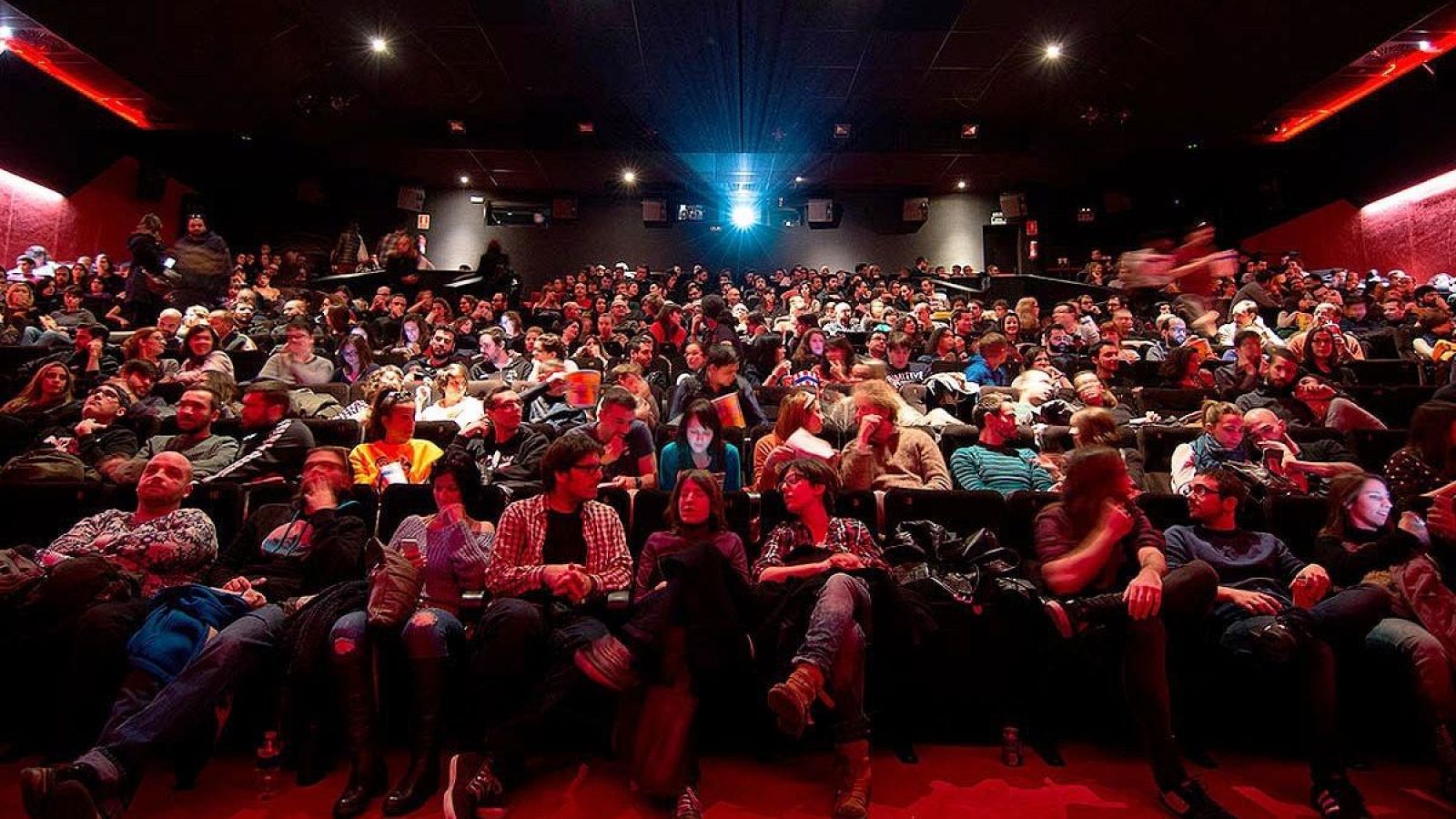 Sala de cine llena, ambiente tenue, multitud expectante en butacas rojas, algunos conversan, otros miran la pantalla.