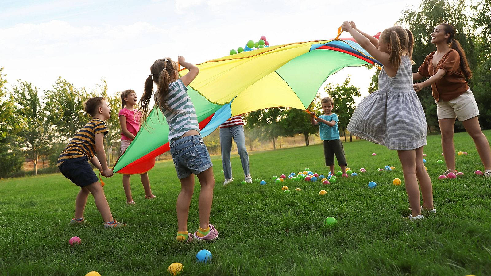 Grupo de niños jugando con un paracaídas de colores en un parque soleado.  Pelotas y una adulta presente.