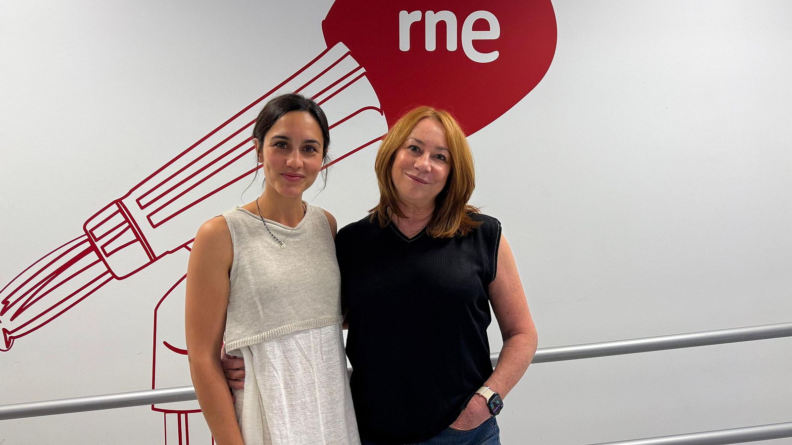 Dos actrices posan juntas frente a una pared con el logo de Radio Nacional de España. Una viste un vestido claro y la otra una camiseta negra y vaqueros.
