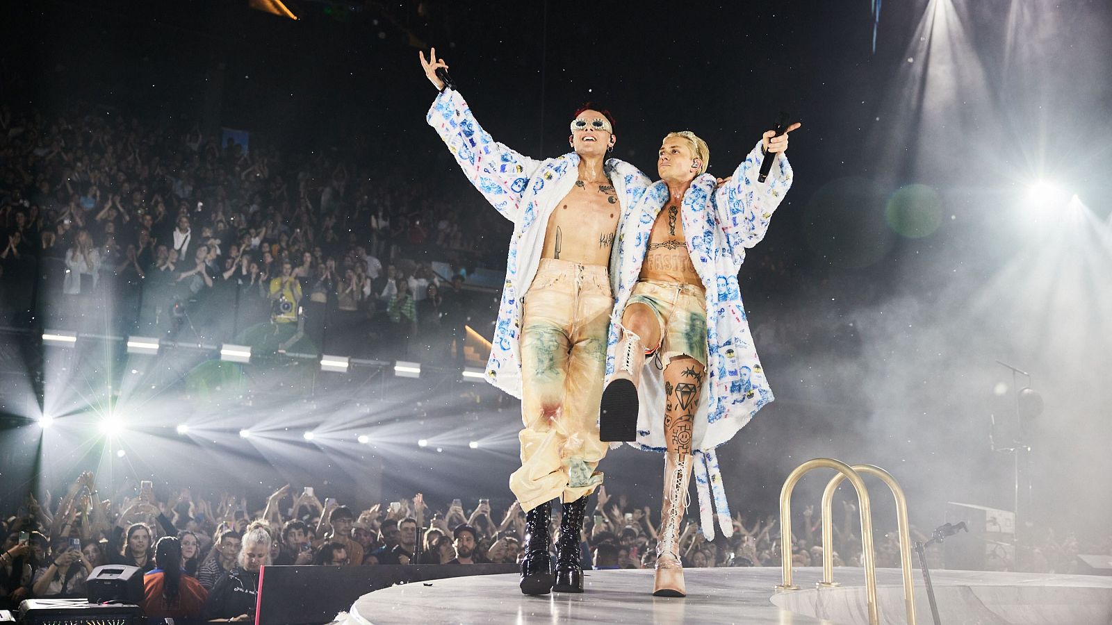 Concierto con dos artistas en escena, vestidos con batas blancas y actuando ante un público. Uno sostiene un micrófono, el otro interactúa con la audiencia.