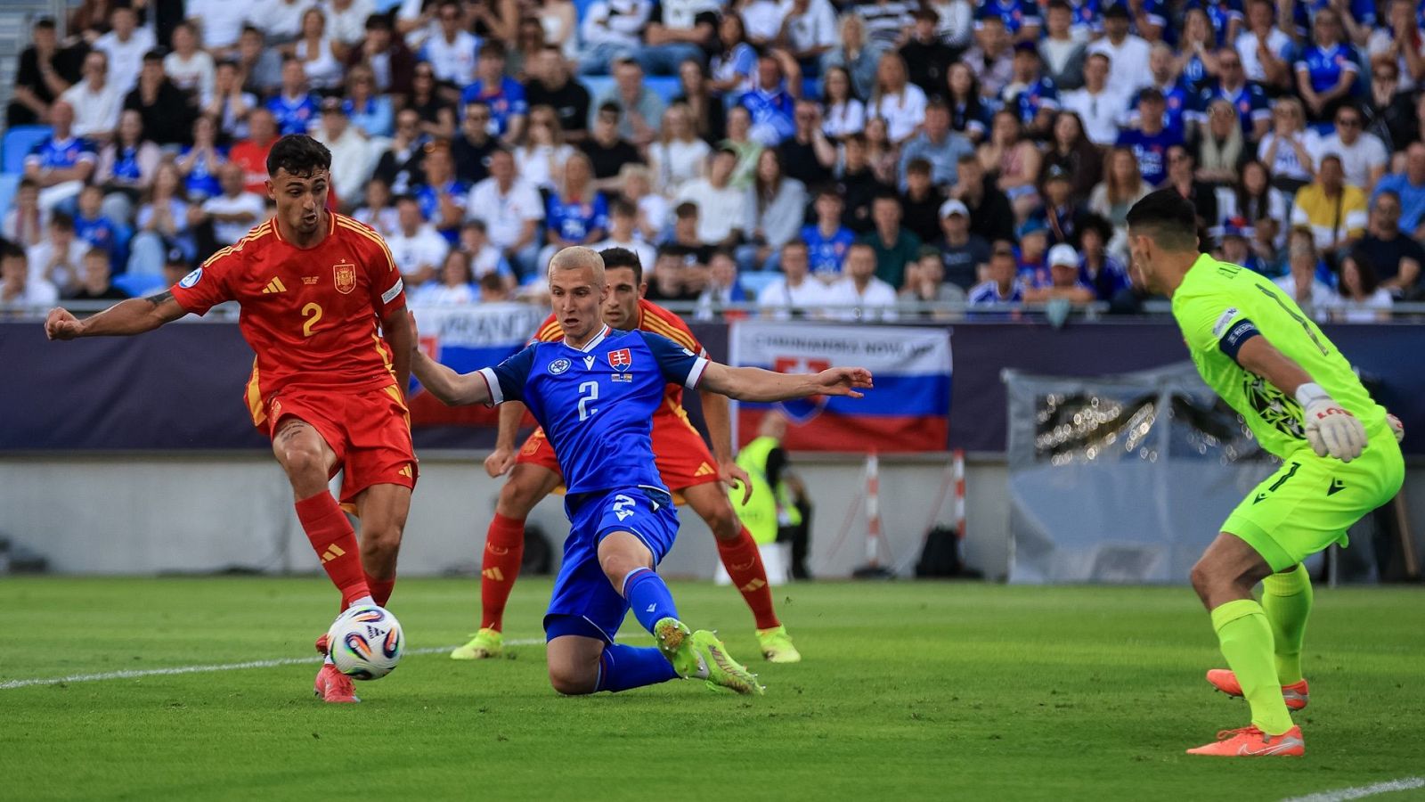 Partido de fútbol: jugador eslovaco (camiseta azul, #7) intenta interceptar el balón mientras un jugador español (camiseta roja, #2) lo conduce. Un portero (verde fluorescente) observa.