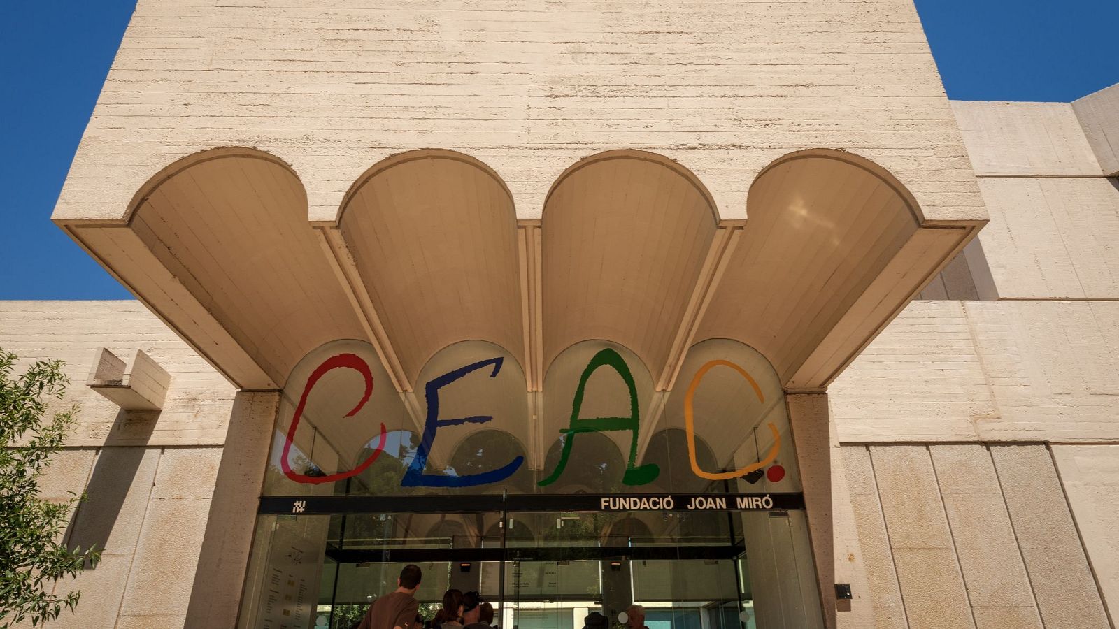 Façana d'un edifici beix clar amb entrada amb arcs i el nom "CEAC" en grans lletres de colors.  Persones entren a l'edifici.