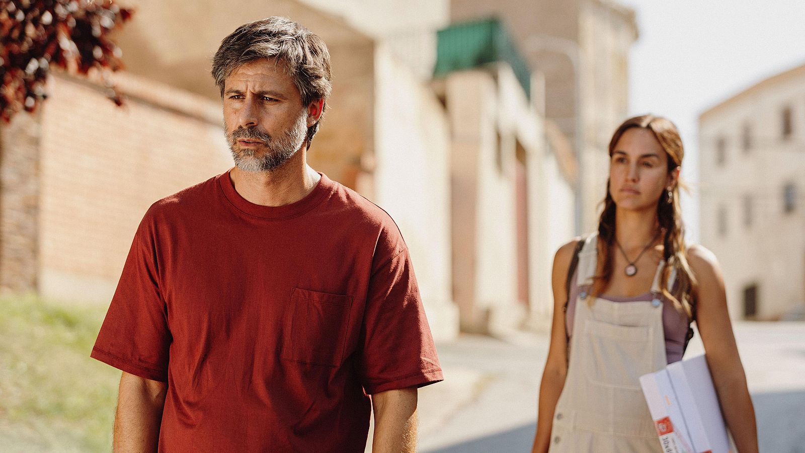 Escena de la película "La buena suerte" con Hugo Silva (camiseta roja) y Megan Montaner (peto beige), caminando por una calle soleada.
