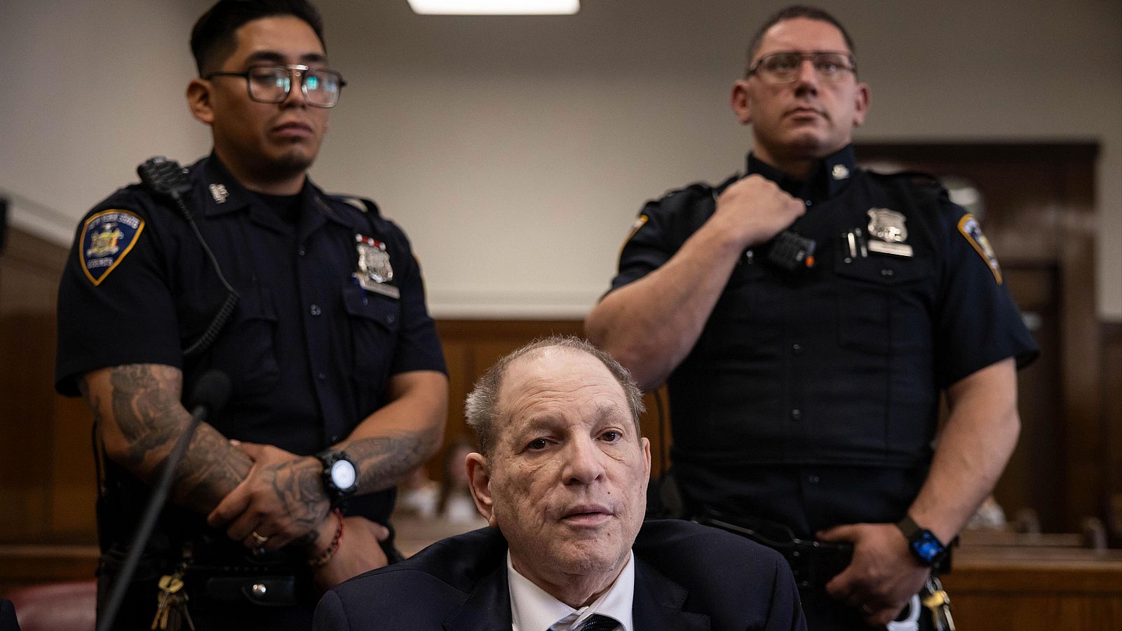 Harvey Weinstein, en juicio en Manhattan, viste traje oscuro y está sentado, custodiado por dos policías de Nueva York, uno con tatuajes visibles.