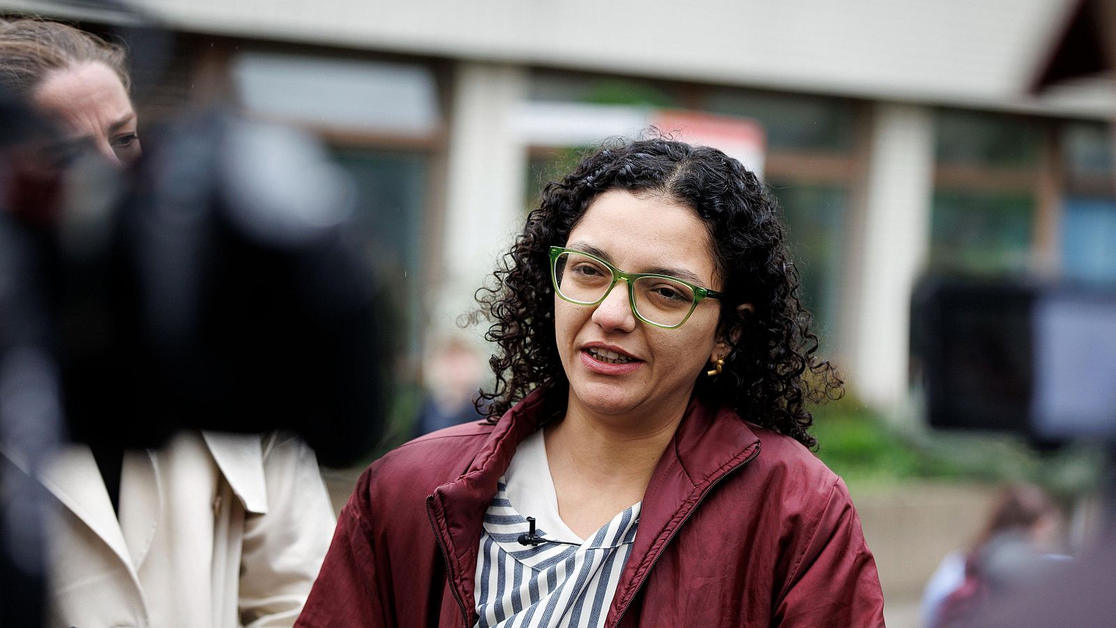 Mujer con chaqueta granate y gafas verdes habla con la prensa frente a un hospital, con un micrófono visible. Parece dar una declaración a los medios.