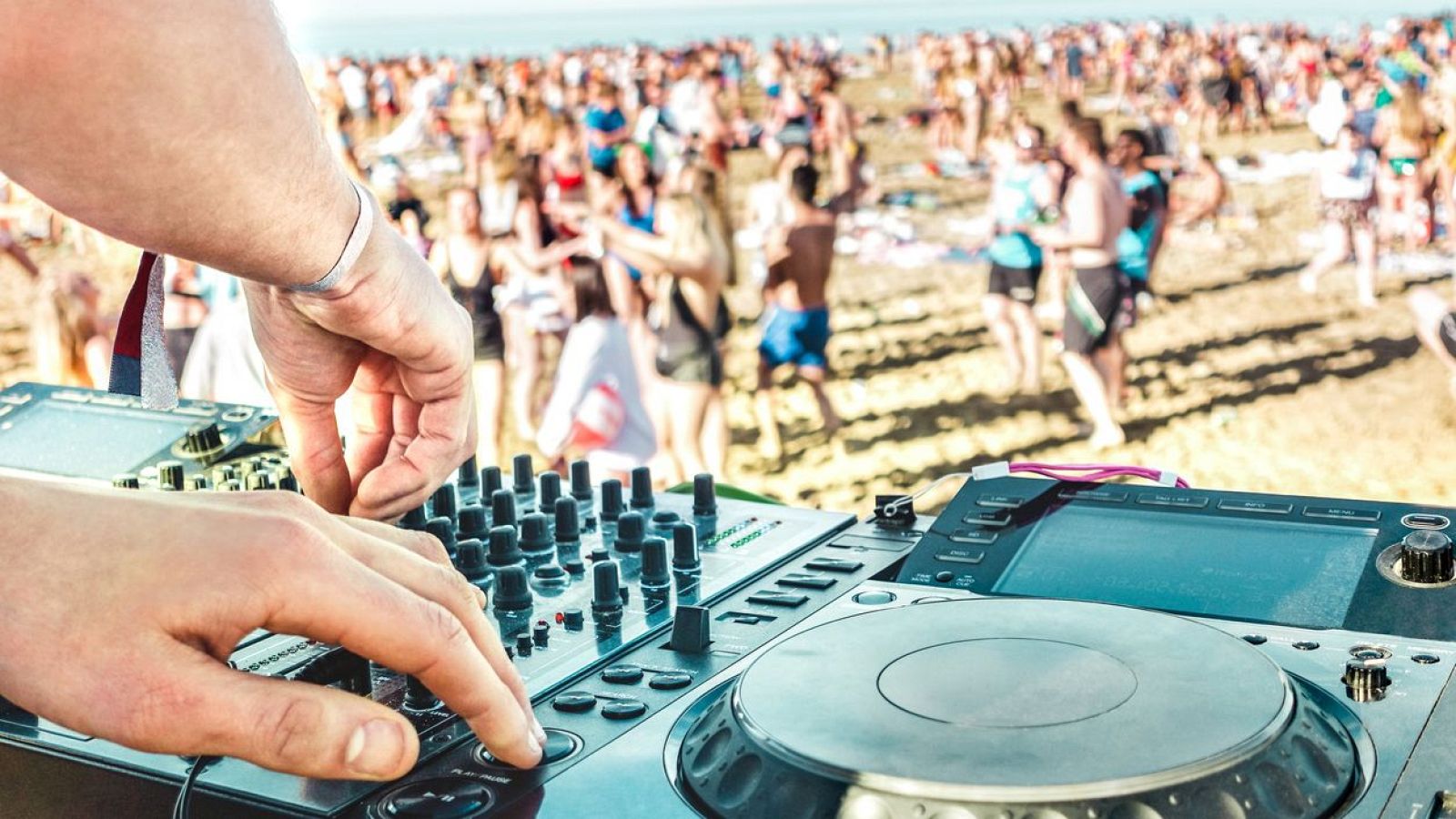 DJ manipulando una mesa de mezclas en un evento al aire libre.  Sus manos sobre los controles, fondo con multitud borrosa bailando en una zona arenosa.