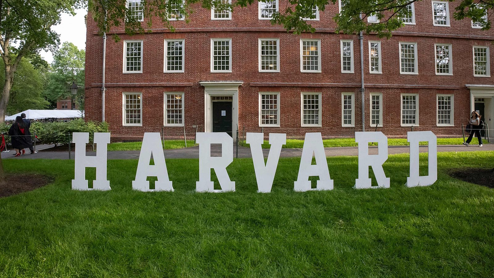 Letras blancas gigantes de "HARVARD" en un césped verde, con un edificio de ladrillo rojo al fondo y personas, una con posible toga, sugiriendo una ceremonia universitaria.