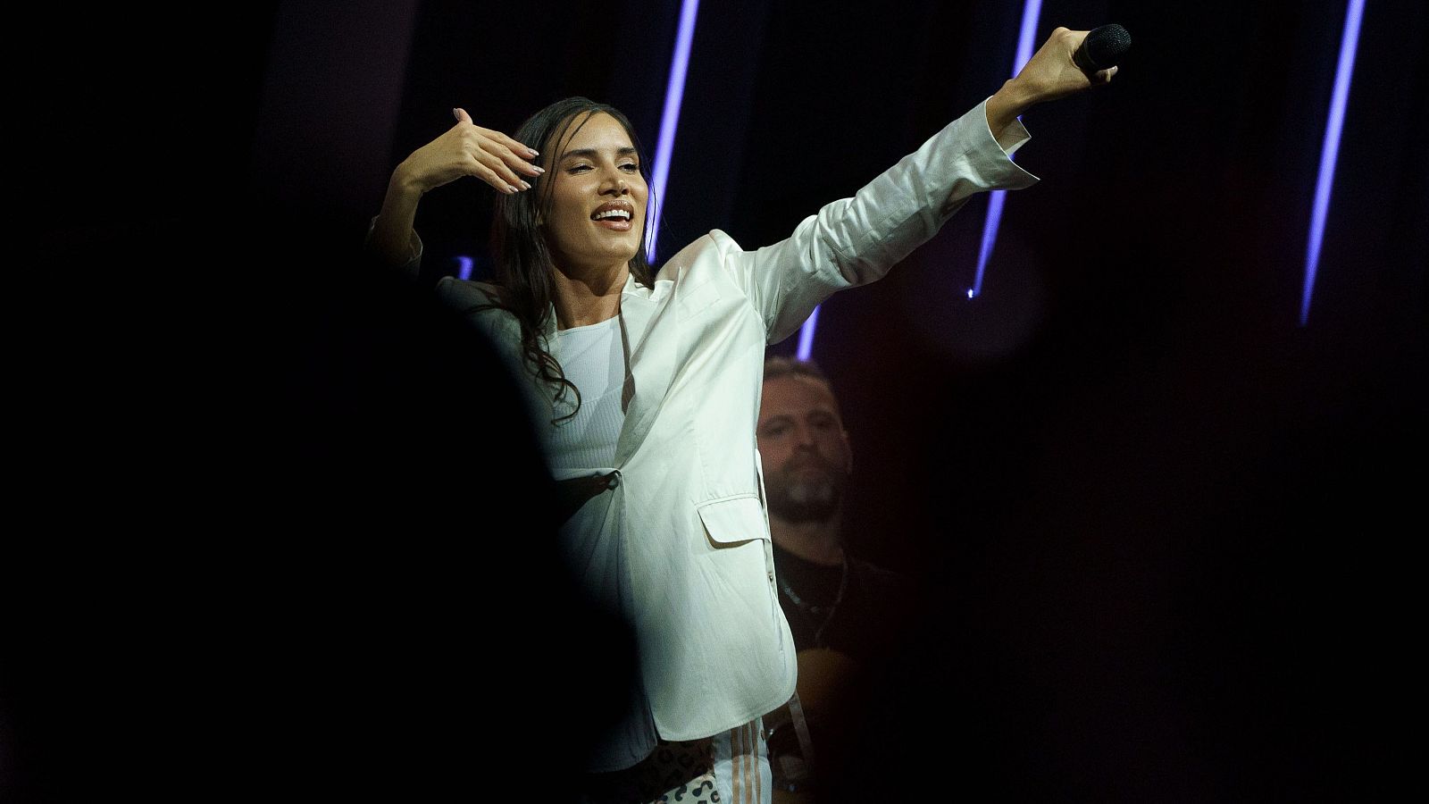 Cantante con americana clara y micrófono en mano, actuando en escenario con luces violetas.  Gestos expresivos y músico en el fondo.