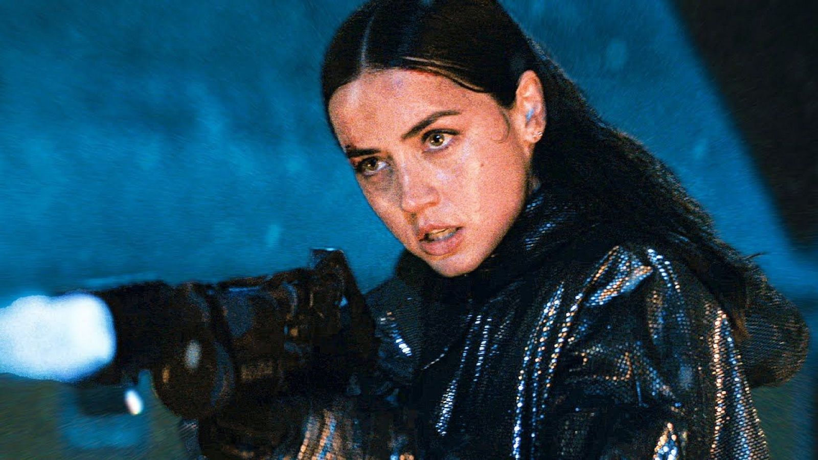 Actriz con expresión seria disparando un arma en escena nocturna; viste chaqueta oscura con brillo. 