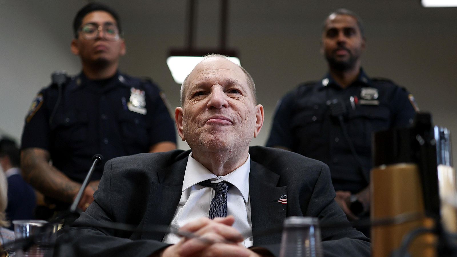 Imagen de Harvey Weinstein en una sala de juicios, con traje oscuro y custodiado por dos policías uniformados.
