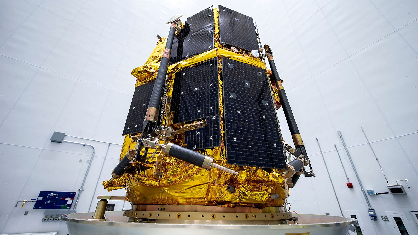 Módulo lunar hexagonal con paneles solares y cubierta térmica dorada, montado en adaptador de cohete dentro de una instalación. Se observan brazos de soporte.