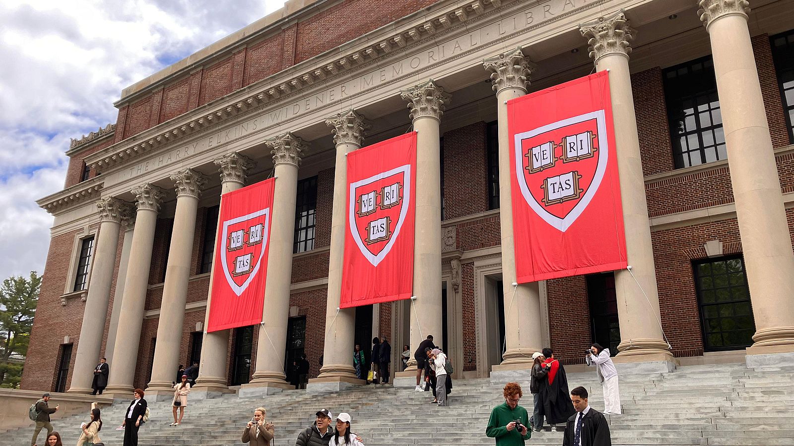 Fachada de la biblioteca Widener de Harvard, con columnas blancas y banderas rojas con el escudo universitario. Personas en las escaleras.