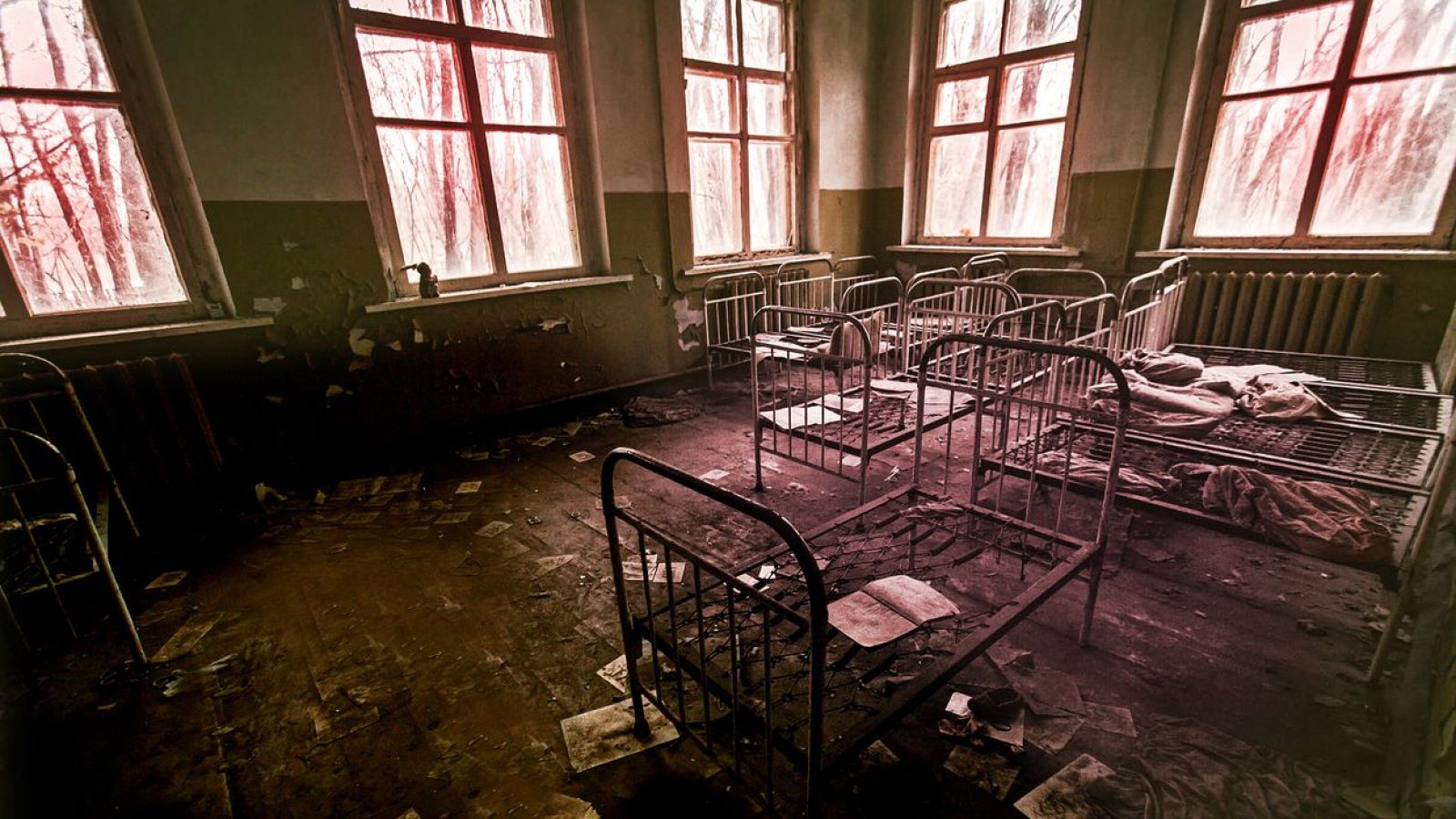 Habitación abandonada de hospital psiquiátrico con suelo sucio, paredes descascarilladas, camas metálicas oxidadas y luz rojiza tenue.  Papeles esparcidos y radiadores antiguos.