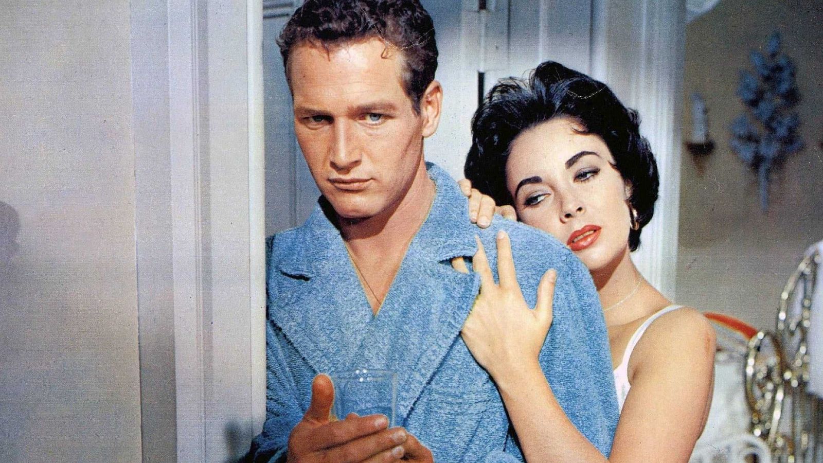 Escena íntima entre Paul Newman y Elizabeth Taylor en un dormitorio. Él, con bata azul, sostiene un vaso; ella, con camiseta clara, lo abraza.