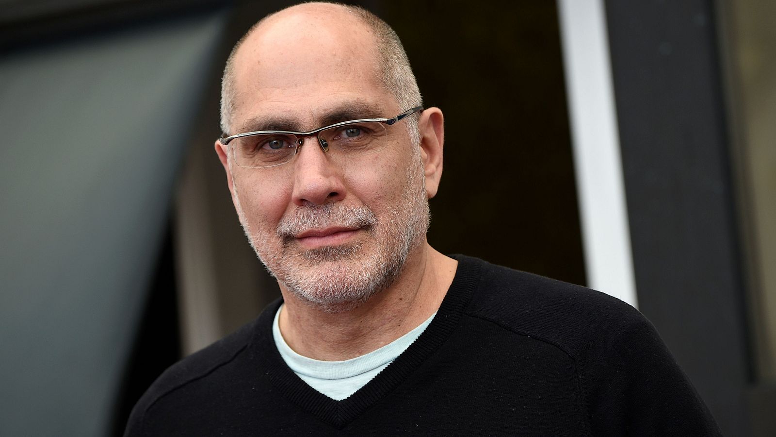 Retrato de Álex de la Iglesia con gafas, cabeza afeitada y barba canosa; viste jersey negro sobre camiseta clara. Fondo oscuro y desenfocado.