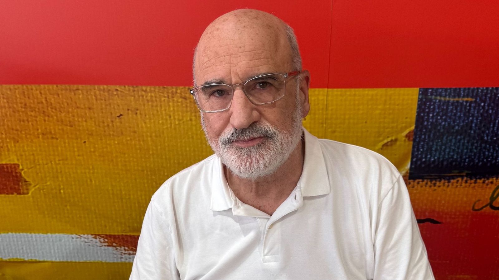 Retrato de hombre mayor con barba blanca, gafas y camisa blanca, sobre fondo de pintura abstracta con rojos, amarillos y azules.