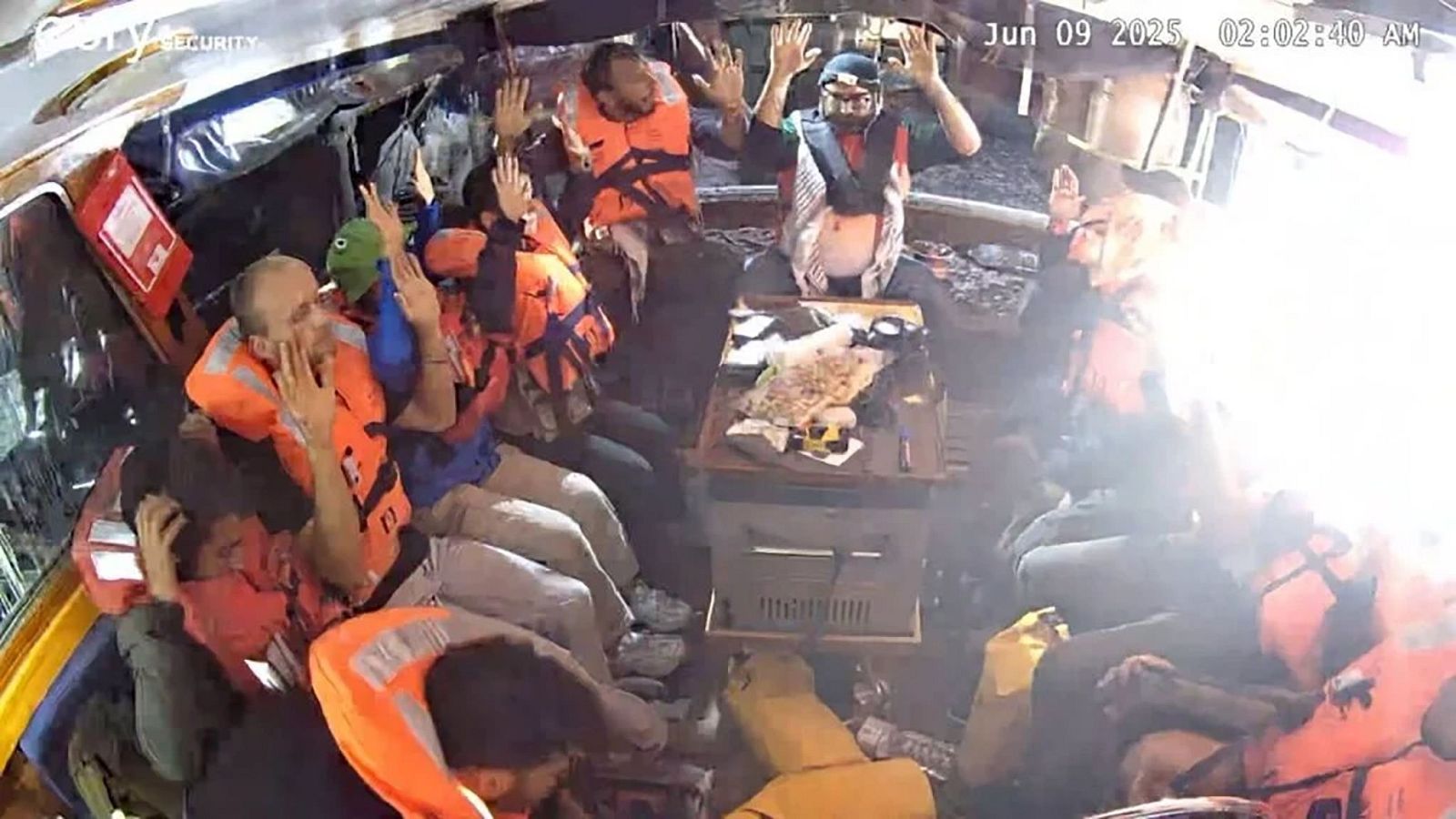 Tripulación de barco, con chalecos salvavidas, algunos con manos levantadas.  Imagen de cámara de seguridad; fecha y hora: 9 de junio de 2025, 02:02:40 AM.