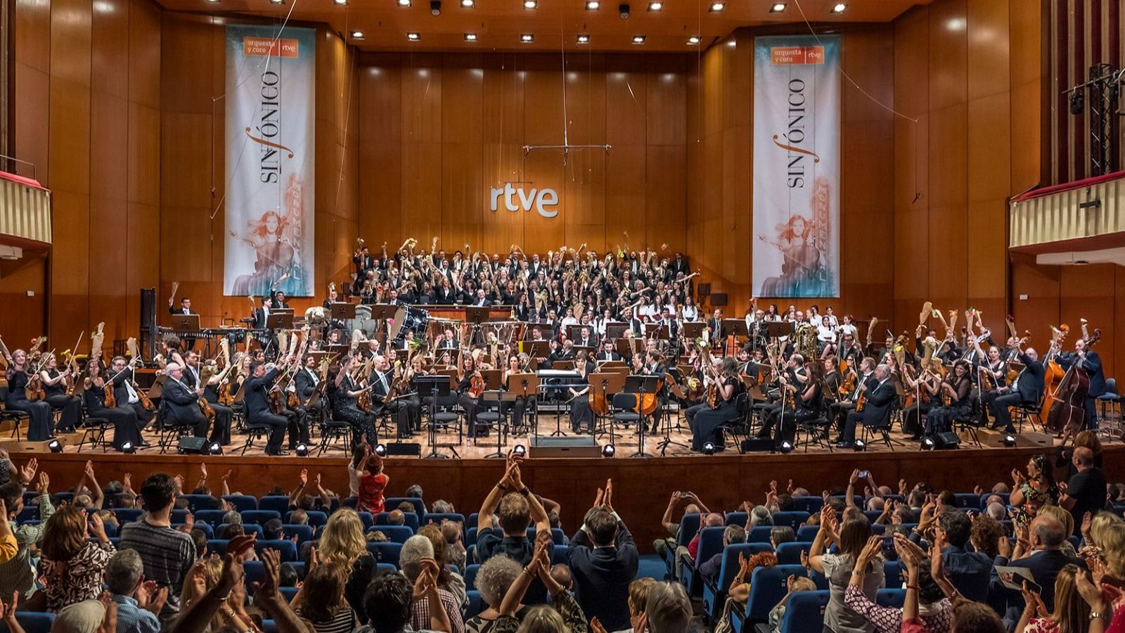 Concierto sinfónico: la Orquesta y Coro de RTVE son ovacionados por un público entusiasmado en un auditorio con asientos azules y paredes de madera. 