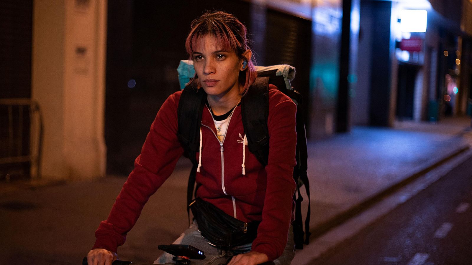 Mujer joven con pelo rosa en bicicleta, sudadera roja y mochila, en calle nocturna. Escena de la película 'Rider'.