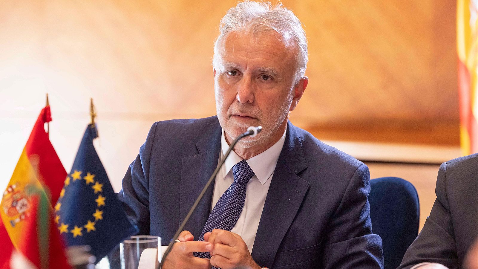 Ministro en traje escuchando atentamente durante un evento oficial.  Banderas de España y la UE al fondo. Micrófono presente.