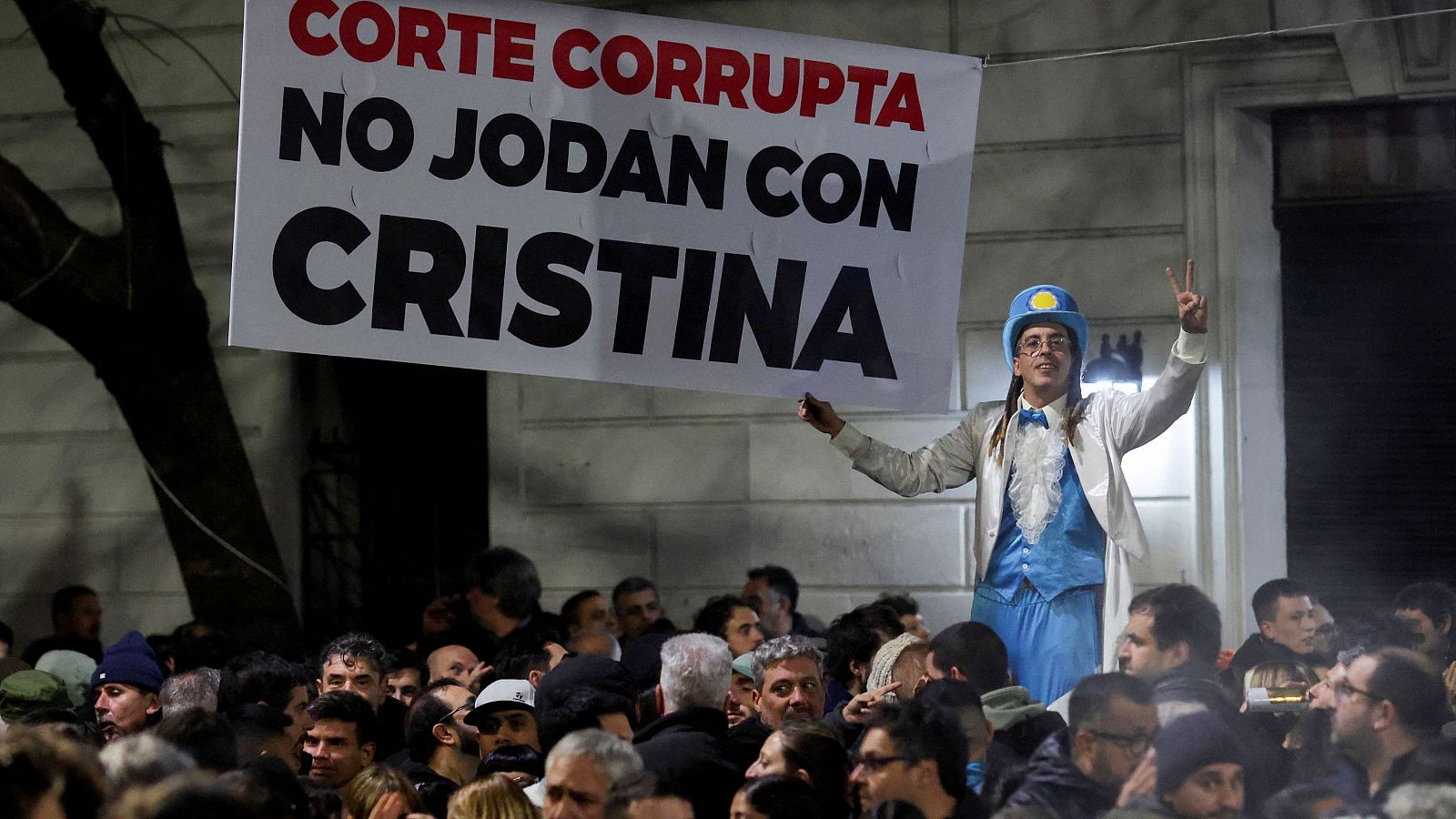 Protesta nocturna en Argentina. Un hombre disfrazado con pancarta "CORTE CORRUPTA NO JODAN CON CRISTINA" y gesto de paz, rodeado de multitud.