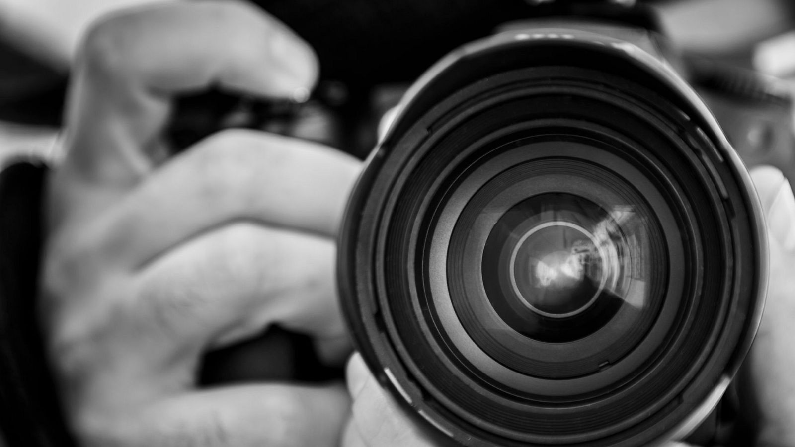 Fotografía en blanco y negro de una cámara con teleobjetivo; el objetivo está enfocado, mostrando círculos concéntricos y un reflejo, mientras que las manos que la sujetan están desenfocadas.