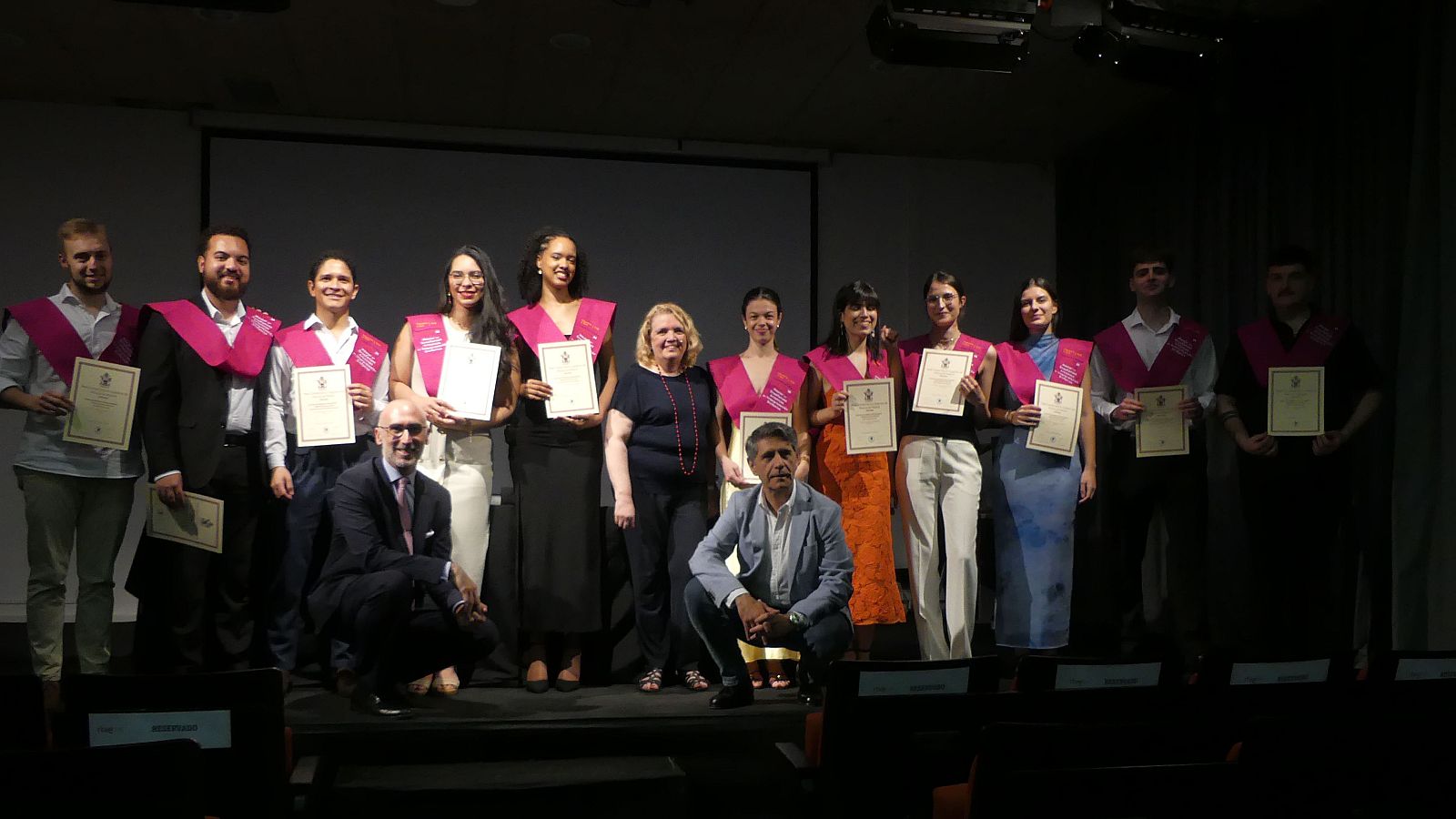 Graduados en traje oscuro y becas rosas posan con sus diplomas tras finalizar el máster.  Presentes profesores o personal.