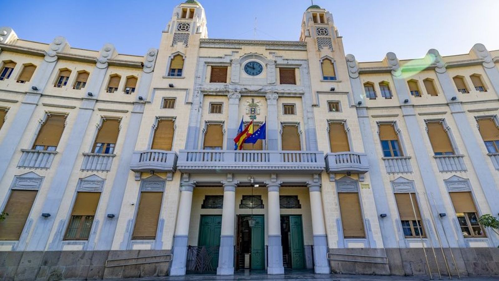 Fachada simétrica de un edificio clásico con dos torres, banderas de España y la UE en un balcón central y rampa de acceso.