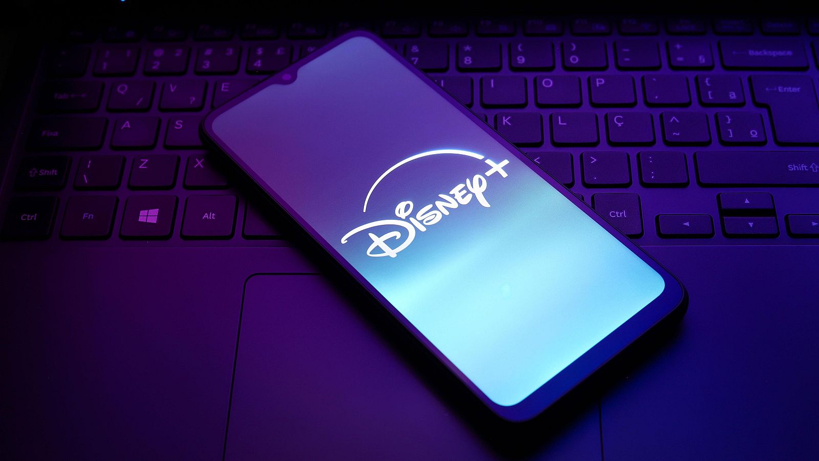 Teléfono móvil mostrando el logo de Disney+ en su pantalla iluminada, apoyado sobre un teclado de portátil con fondo oscuro. Iluminación púrpura y azulada.
